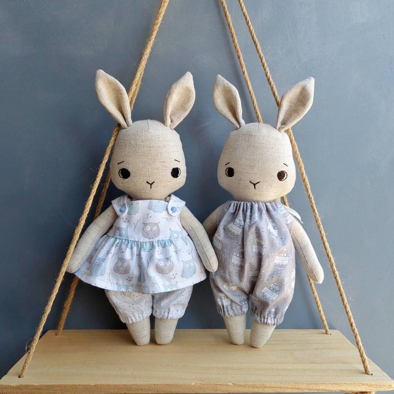 May include: Two handmade plush bunny dolls with embroidered faces. One doll is wearing a blue and white dress with an owl print, the other is wearing a gray and white dress with a floral print.