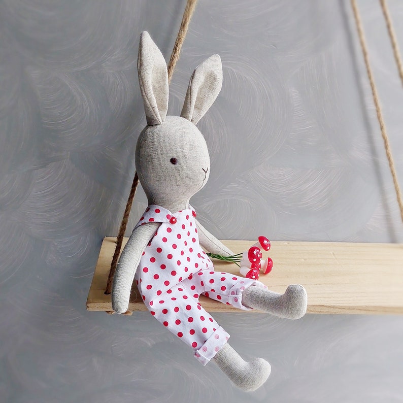 May include: A gray linen bunny doll wearing a white and red polka dot dress. The doll is sitting on a wooden swing set.