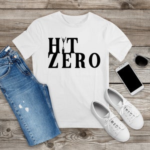 May include: White t-shirt with black text that reads "HIT ZERO" with a stick figure jumping over the word "ZERO". The shirt is laid out on a wooden surface with blue jeans, white sneakers, a smartphone, and sunglasses.
