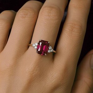 May include: A gold ring featuring a large, rectangular, deep red gemstone as the centerpiece. The ring is accented with smaller, clear gemstones on either side of the main stone. The ring is set on a hand.