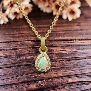 May include: A gold-colored necklace with a pear-shaped pendant. The pendant features a central opal stone surrounded by small, clear gemstones. The necklace is displayed on a wooden surface.
