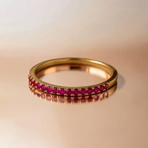 Könnte beinhalten: Ein dünner Goldring mit einer Reihe kleiner, runder, leuchtend pink-roter Edelsteine, die entlang der vorderen Hälfte des Bandes gefasst sind. Das Metall hat eine polierte Oberfläche und der Ring ist so positioniert, dass seine Form sichtbar ist.