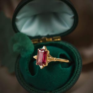 Könnte beinhalten: Ein goldener Ring mit einem rechteckigen, tiefroten Edelstein und zwei kleineren weißen Edelsteinen. Der Ring wird in einer offenen, dunkelgrünen Samt-Schmuckschatulle präsentiert. Das Design des Rings ist von Vintage inspiriert.