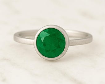 May include: A silver ring featuring a large, round, faceted emerald gemstone. The bright green stone is set in a simple bezel setting, creating a classic and elegant look. The ring's band is a thin, polished silver.