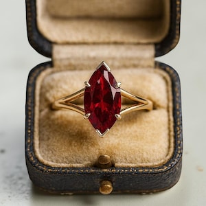 Può includere: Un anello d'oro con una gemma rosso scuro taglio marquise. L'anello è presentato in un portagioie blu scuro in stile vintage con interno beige. La gemma rossa è il punto focale, mostrando il suo colore e taglio.