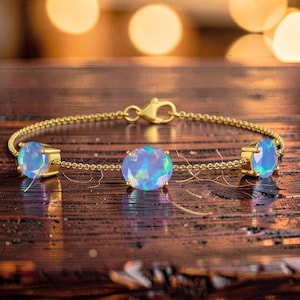 May include: A gold bracelet with three oval, iridescent opal stones. The bracelet has a delicate chain and a clasp closure. The opals display a range of colors, including blue, green, and purple, set against a warm wooden background.