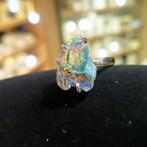 May include: A silver ring featuring a large, irregularly shaped gemstone with a vibrant display of colors, including blues, greens, and oranges. The gemstone has a rough, textured surface, and the ring band is a simple, polished silver.