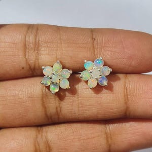 May include: A pair of flower-shaped stud earrings. Each earring has a cluster of small, iridescent opal stones set in silver metal. The opals display white, green, and blue colours. The earrings are a delicate floral design.