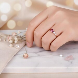 May include: A rose gold ring featuring a pear-shaped pink gemstone, flanked by two smaller white gemstones. The ring is set on a marble surface, with small flowers and pearls in the background.