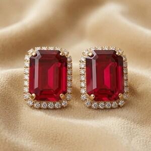 May include: A pair of rectangular earrings featuring large, deep red, emerald-cut centre stones. Each central red stone is surrounded by a halo of small, clear, round-cut white stones set in a gold-toned metal frame with four corner prongs.