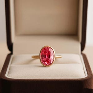 May include: A gold ring featuring a large, oval-cut, vibrant pinkish-red gemstone set in a smooth, polished bezel setting. The band is thin and circular, crafted from a warm-toned yellow metal that complements the central stone's facets.
