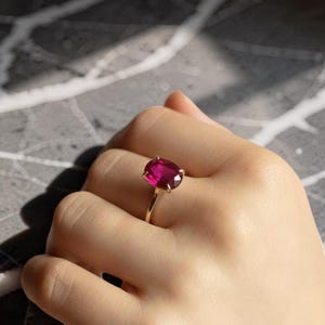 May include: A gold ring with a large, oval-cut ruby gemstone. The ruby is a vibrant red, set in a classic four-prong setting. The ring's band is a simple, polished gold.