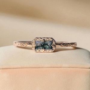 Puede incluir: Anillo de oro rosa con una piedra de ágata musgosa rectangular. El anillo tiene un engaste ornamentado y una banda con un sutil patrón floral. El ágata musgosa muestra inclusiones verdes y blancas, creando un aspecto natural y único.