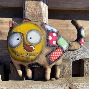 May include: A whimsical, handmade cat plushie with a yellow face, large white eyes, and a brown tongue. The cat's body is tan with colorful patchwork details in red, green, and blue. The cat is standing on a wooden surface.