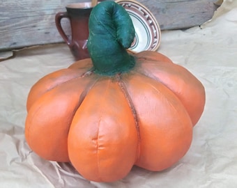 Può includere: Una zucca arancione fatta a mano con uno stelo verde, realizzata in tessuto e dipinta con una finitura opaca. La zucca è appoggiata su una superficie di carta marrone.