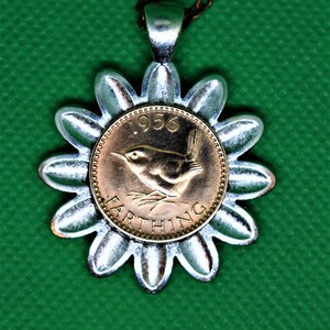 May include: A silver flower-shaped pendant with a 1956 Farthing coin at its core, depicting a bird. The pendant is suspended from a thin, brown chain. The background is a solid green colour.
