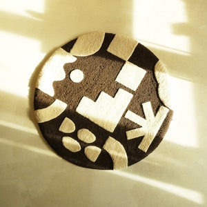 May include: A circular rug featuring a dark brown base with a contrasting cream-colored abstract pattern. The design consists of various geometric shapes, including a stepped block, a small circle, and several curved and angular lines.