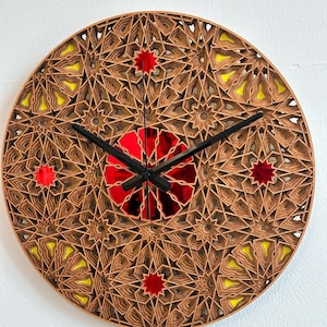 Op de afbeelding: Een ronde houten klok met een geometrisch ontwerp met rode en gele accenten. De wijzerplaat van de klok is gemaakt van gelaagd hout en heeft een gespiegelde kern.