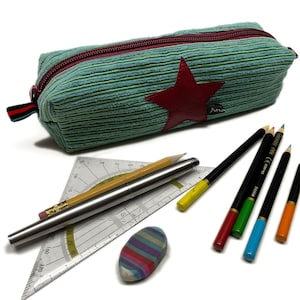 Op de afbeelding: Een groen-rood gestreepte etui met een rode ster en de naam "Ana" erop geborduurd. De etui is open en bevat potloden, een liniaal en een gum.