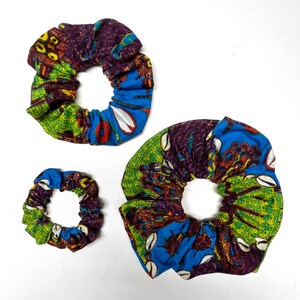Puede incluir: Tres coleteros de tela coloridos de diferentes tamaños. Los coleteros presentan una vibrante mezcla de patrones azules, verdes, morados y amarillos. La tela tiene un diseño audaz de inspiración africana, con acentos en forma de concha.