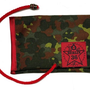May include: A camouflage pattern wallet with red trim and a red drawstring. The wallet has a red patch with the text "TRASH BERLIN 36 UP" and a star graphic. The drawstring is attached with a metal skull-shaped bead.