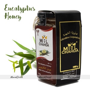 May include: A glass jar of honey with a black label. The label has a gold butterfly logo and the text "MIEL CHAHDA" in gold lettering. The jar is labeled "Eucalyptus Honey" and "1 kg". The jar is sitting on a white surface with a eucalyptus branch in the background.