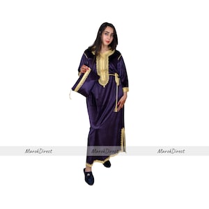 Puede incluir: Un vestido largo de terciopelo morado con bordados y ribetes dorados. El vestido tiene un cinturón y un borla en la cintura. El vestido lo lleva una persona de pie sobre un fondo blanco.