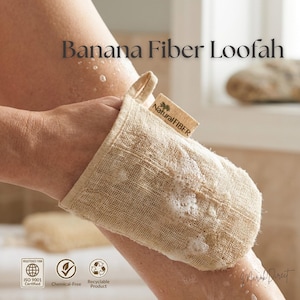 May include: A beige banana fibre loofah mitt with the text "Banana Fiber Loofah" and "Natural FIBER". The mitt is being used on a person's leg. The loofah is made of a woven material and has a small loop for hanging.