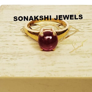 Peut inclure: Une bague en or avec une grande pierre précieuse ronde de couleur rose foncé. La bague est présentée dans une boîte blanche avec le texte "SONAKSHI JEWELS" sur le dessus.