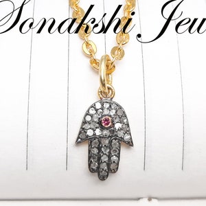 May include: A gold chain necklace with a hamsa hand pendant. The pendant is set with small diamonds and a single red gemstone in the center. The hamsa hand is a symbol of protection and good luck.