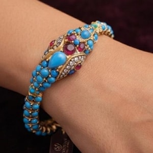 Könnte beinhalten: Ein goldfarbenes Armband mit einem schlangenförmigen Kopf, der mit runden Türkisperlen, facettierten roten Steinen und kleinen klaren Kristallen besetzt ist. Das Band besteht aus Reihen kleiner Türkisperlen und ein kleines rechteckiges Etikett hängt am Verschluss.