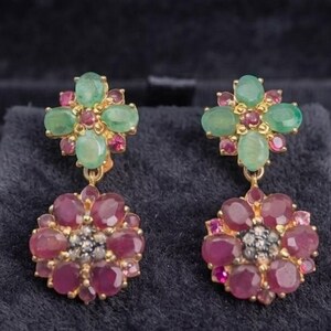 May include: A pair of gold earrings featuring a top cluster of four oval green emeralds and small red rubies. The dangling portion consists of a circular arrangement of deep red rubies surrounding a central cluster of tiny dark grey stones.