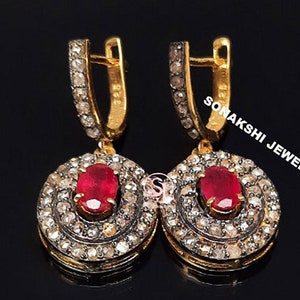 May include: A pair of gold earrings featuring a red ruby gemstone set within a halo of small diamonds. The earrings have a hoop design and are marked "SONAKSHI JEWELS".