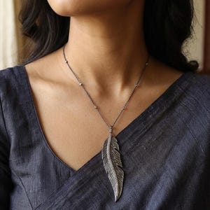 May include: A dark metal necklace featuring a long, textured feather pendant with a central row of small, clear stones. The chain is thin and dark, accented with evenly spaced, tiny, clear stones along its length, resting against a dark fabric.