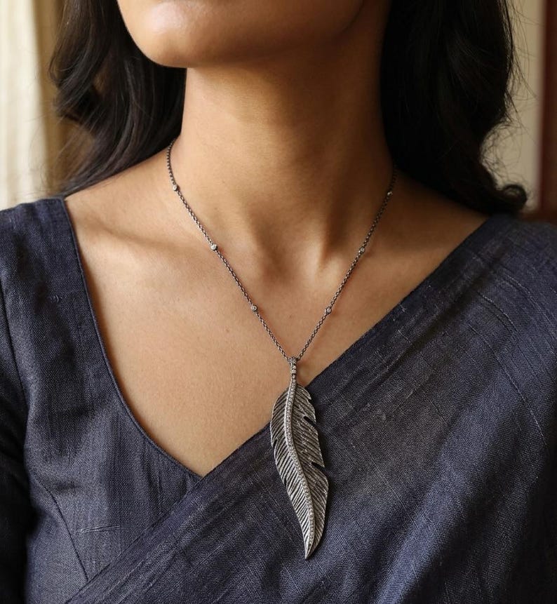 May include: A dark metal necklace featuring a long, textured feather pendant with a central row of small, clear stones. The chain is thin and dark, accented with evenly spaced, tiny, clear stones along its length, resting against a dark fabric.