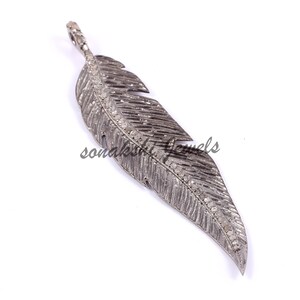 May include: A dark grey metal feather pendant featuring a textured surface and a central spine encrusted with small, clear pav&eacute;-set stones. The piece includes a small integrated bail at the top for suspension, with the text sonakshi jewels visible.