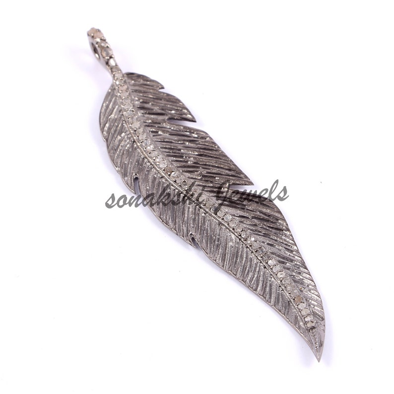 May include: A dark grey metal feather pendant featuring a textured surface and a central spine encrusted with small, clear pav&eacute;-set stones. The piece includes a small integrated bail at the top for suspension, with the text sonakshi jewels visible.