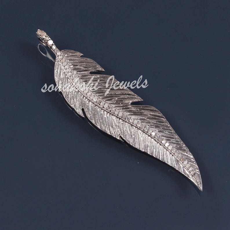 May include: A silver feather pendant with pave set diamonds. The feather is detailed with lines and has a loop at the top for attaching to a chain. The pendant is on a blue background with the text 'sonashi jewels' visible.