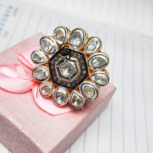 May include: A silver ring with a large, round, flower-shaped design. The ring is set with many small, clear stones. The center of the flower is a hexagonal shape with a larger, clear stone.