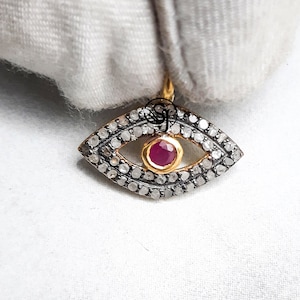May include: A gold-toned pendant in the shape of an eye, encrusted with small, clear stones around the perimeter. The center features a vibrant red gemstone set in a gold bezel. The pendant is displayed against a white background.