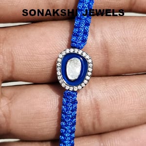 May include: A blue woven bracelet with a central charm. The charm features a white stone set in a silver-colored frame, surrounded by a blue enamel border and small white stones. The text "SONAKSHI JEWELS" is visible.
