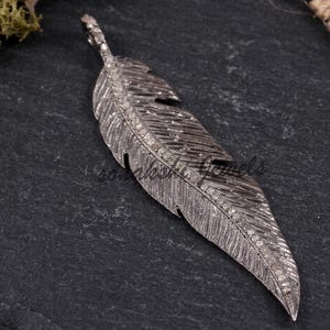 May include: A dark grey metal feather pendant featuring a central spine encrusted with small, clear, sparkling stones. The surface of the feather has detailed, etched lines mimicking natural plumage, with a small loop at the top for attachment.