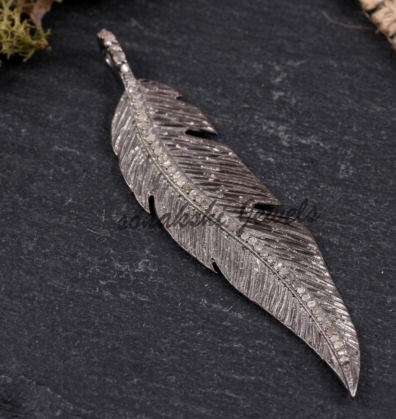 May include: A dark grey metal feather pendant featuring a central spine encrusted with small, clear, sparkling stones. The surface of the feather has detailed, etched lines mimicking natural plumage, with a small loop at the top for attachment.
