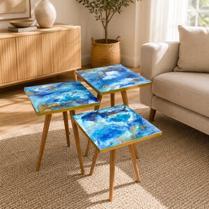 Pode incluir: Três mesas laterais quadradas com um design abstrato azul e dourado no tampo. As mesas têm pernas de madeira castanho-claro e uma borda dourada. Estão dispostas sobre um tapete bege.