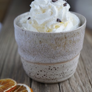 Könnte beinhalten: Eine weiße Keramiktasse mit gesprenkeltem Glase, gefüllt mit Schlagsahne und Schokoladenstreuseln. Die Tasse steht auf einer Holzoberfläche mit einer getrockneten Orangenscheibe daneben.