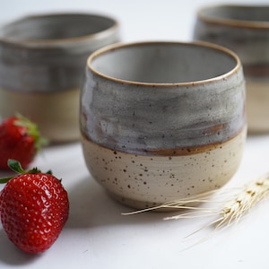Op de afbeelding: Drie keramische mokken met een gespikkeld glazuur in tinten grijs en beige. De mokken staan op een wit oppervlak met twee rode aardbeien en een tarwehalm.