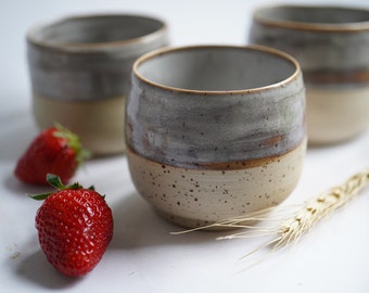 May include: Three ceramic mugs with a speckled glaze in shades of gray and beige. The mugs are sitting on a white surface with two red strawberries and a stalk of wheat.