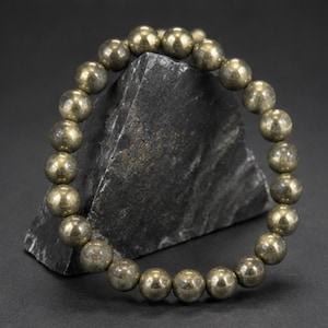 May include: A circular bracelet composed of polished, spherical pyrite beads with a metallic, brass-like luster. The beads are uniform in size and strung together to form a continuous loop, displaying a reflective, golden-grey surface texture.