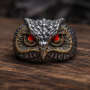 Peut inclure: Une bague en métal argenté en forme de tête de hibou avec des détails de plumes complexes. Le hibou présente des yeux circulaires rouge vif et des accents dorés autour des orbites, avec une finition sombre oxydée dans les zones en creux.