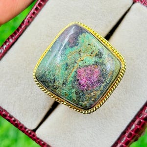 Puede incluir: Anillo cuadrado con una gran piedra preciosa multicolor. La piedra muestra tonos verdes, morados y negros, engastada en un marco dorado con textura de cuerda. El anillo se presenta en una caja de joyería roja.